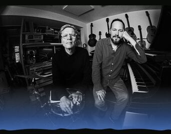 Det sort-hvide foto af to musikere, der står sammen foran flere musikinstrumenter.