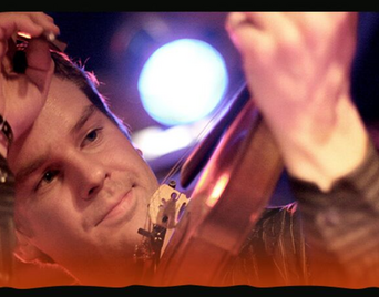 Foto af en musiker med et strygeinstrument i hånden og scenelys i baggrunden.