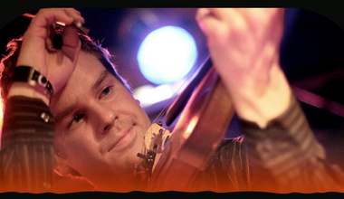 Foto af en musiker med et strygeinstrument i hånden og scenelys i baggrunden.